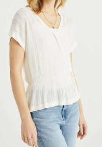 Top bianco lavorato a maglia con maniche corte, scollatura a V e vita elastica. Presenta righe verticali e un dettaglio con lacci su un lato. Abbinato a jeans azzurri.