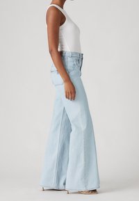 Woman in light blue high-waist wide-leg jeans, white sleeveless top, and beige heeled sandals standing against a plain gray background.