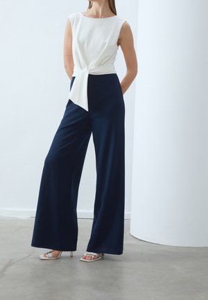 Vrouw draagt een mouwloos wit topje met zijstrik, marineblauwe broek met wijde pijpen en zilveren sandalen met hoge hakken, staand op een betonnen vloer.
