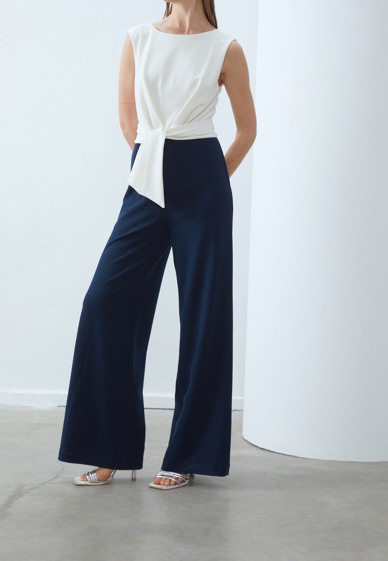 Femme portant un haut blanc sans manches avec un lien sur le côté, un pantalon large bleu marine et des sandales à talons hauts argentées, debout sur un sol en béton.