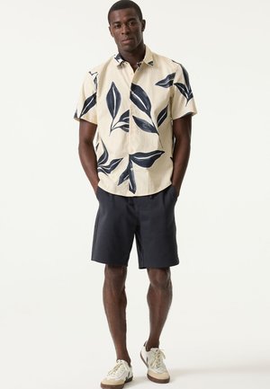 Kurzärmeliges Hemd in Creme mit marineblauem Blättermuster; kombiniert mit dunklen Shorts und weißen Sneakers mit hellbraunen Akzenten.