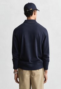 Marineblauer Strickpullover mit langen Ärmeln und Polo-Kragen, kombiniert mit beige Hose. Das Modell trägt eine passende marineblaue Mütze.