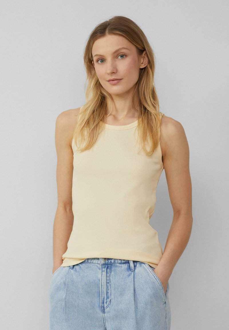 Lichtgele geribde tanktop met een ronde halslijn. Het model draagt lichtblauwe high-waisted jeans, met handen in de zakken, tegen een neutrale achtergrond.