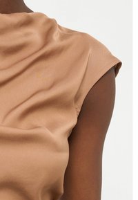 Κοντινό πλάνο στο άνω μέρος του χεριού και τον ώμο ενός ατόμου που φοράει λείο, ανοιχτό καφέ αμάνικο τοπ, σε απλό φόντο.