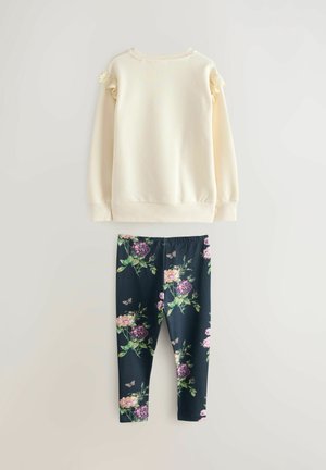 Tricou crem deschis cu accente din dantelă pe mâneci, asortat cu colanți bleumarin cu un model floral în tonuri de roz și violet.