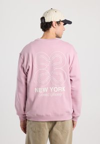Felpa rosa con una vestibilità rilassata e maniche oversize. Sul retro c'è una grafica bianca di forme intrecciate accompagnata dal testo "NEW YORK Tennis Society".