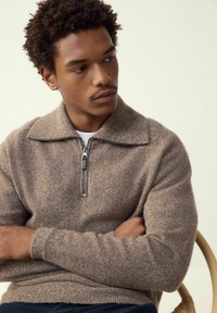 Pruun, kootud pullover kõrge kraega ja lühikese tõmblukuga. Iseloomulik tekstureeritud kangas ning soonikkoes varrukad ja allserv. Modell istub käed rüpes.