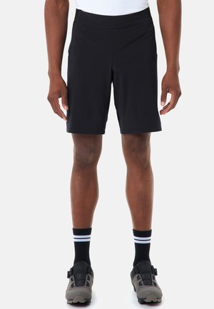 Persoon draagt zwarte sportkorte broek, zwarte sokken met witte strepen en grijze en zwarte fietsschoenen, staand tegen een effen achtergrond.