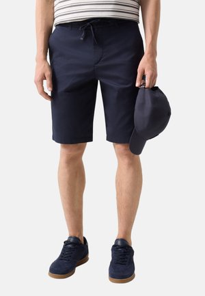 Man draagt marineblauwe shorts, gestreept shirt, marineblauwe sneakers, houdt een bijpassende marineblauwe pet naast zich, staat op een witte achtergrond.
