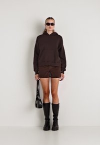 Hanorac cu glugă maro, cu buzunar frontal, asortat cu pantaloni scurți maro. Modelul poartă cizme negre până la genunchi și ochelari de soare, ținând o geantă neagră.