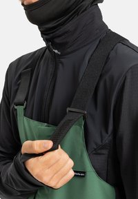 Man past het zwarte elastische suspenderband aan op een groene schort die over een zwart jack met rits en duimgaten wordt gedragen.