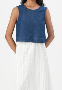 Denimkort topp med ärmlös design, blå färg, rund halsringning och synlig sömnad. Bärs över en vit, mönstrad klänning.