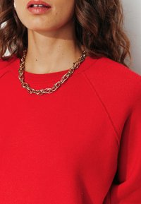 Pull en tricot rouge avec une texture côtelée et des manches raglan, associé à un collier en chaîne dorée épaisse mettant en valeur des maillons ronds.