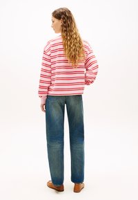 Φούτερ με κόκκινες και λευκές ρίγες, σε άνετη εφαρμογή, σε συνδυασμό με ξεβαμμένα μπλε τζιν και καφέ παπούτσια. Τα μαλλιά είναι χτενισμένα σε χαλαρούς κυματισμούς.