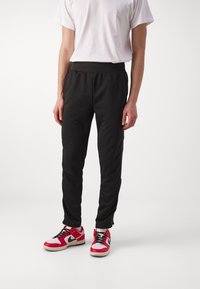 Zwarte sportieve broek met taps toelopende pasvorm, elastische tailleband en zijstrepen. Gecombineerd met rood-witte sneakers met zwarte accenten.