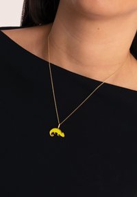 Collier en or avec un pendentif en forme de caméléon jaune et accents noirs, reposant sur un fond en tissu sombre.