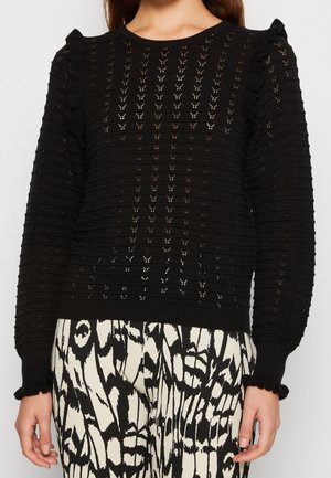 Pull noir en tricot avec un motif texturé, des poignets côtelés et des accents d'épaules volantés. Associé à un pantalon blanc et noir à motifs.