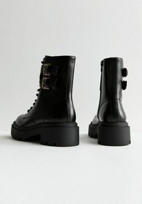 Botas de cuero negro hasta el tobillo con suelas gruesas, con dos hebillas decorativas, parte delantera con cordones y cierre de cremallera en la parte posterior.