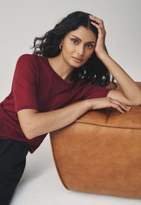 Bordeauxrød kortærmet top med tekstureret finish, kombineret med en tanfarvet læderpuf. Glat overflade og afrundet form er fremtrædende.