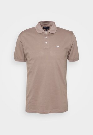 Polo beige à manches courtes avec trois boutons blancs et un petit logo blanc sur le côté gauche de la poitrine sur fond uni.