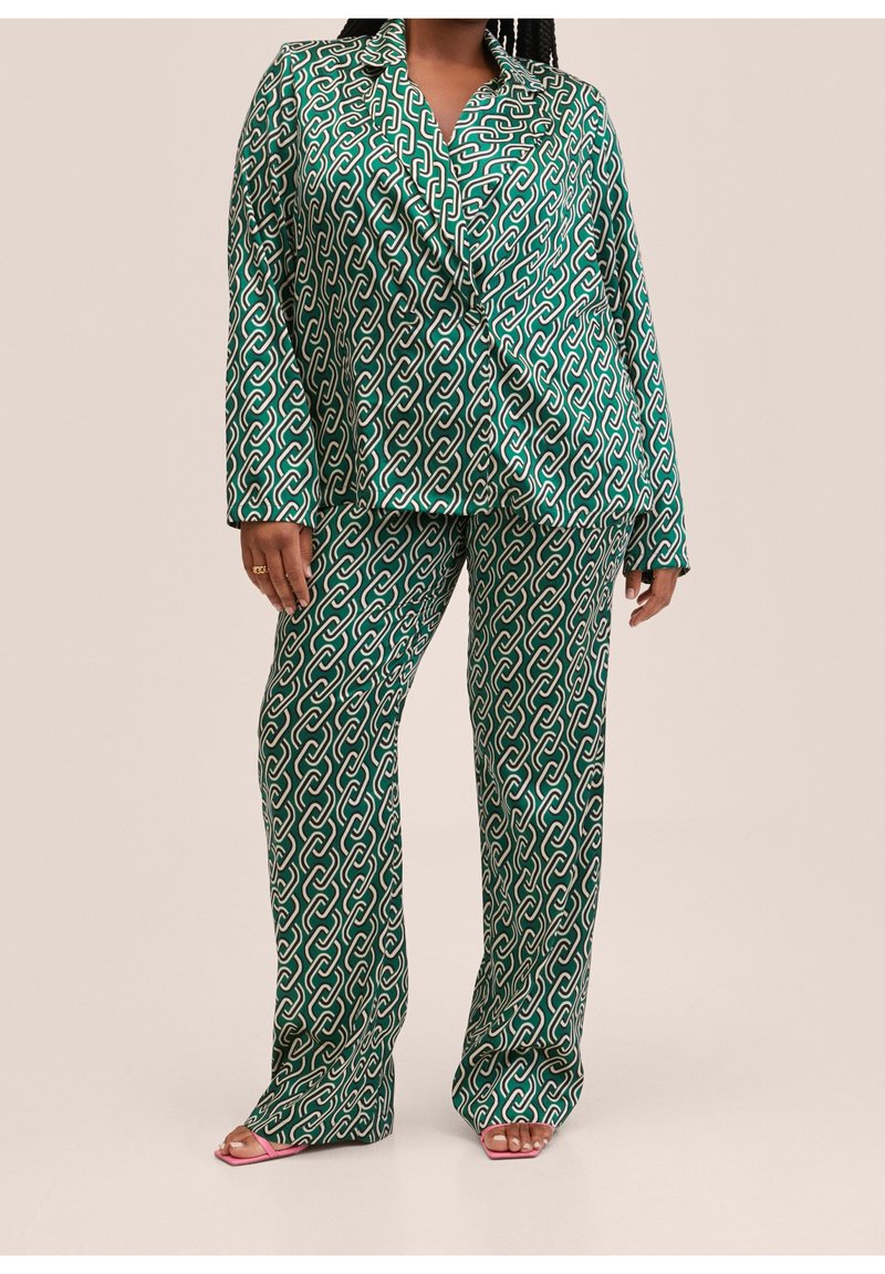 Femme portant un tailleur deux-pièces vert à motif de chaîne entrelacée et des talons ouverts roses, debout devant un fond uni.
