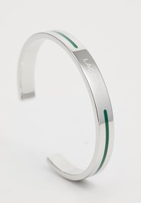 Ezüst színű fémből készült csuklópánt, sima felülettel, a "LACOSTE" felirattal gravírozva, és két zöld díszcsíkkal a külső felületen.