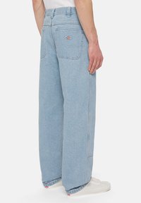 Jasnoniebieskie szerokie dżinsy noszone przez osobę stojącą, ukazujące tylne kieszenie i detale bocznych szwów, w połączeniu z białymi trampkami.