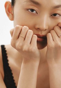 Anillos de oro y plata en los dedos, primer plano de piel suave. Modelo con fondo neutro, vistiendo una blusa negra, manos cerca de la cara.