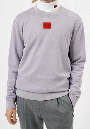 Osoba ubrana w jasnoszarą bluzę z okrągłym dekoltem z czerwonym naszywką "HUGO" na piersi, białą golf z pasującym czerwonym logo oraz szare spodnie.