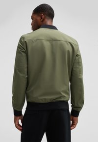 Olijfgroene bomberjack met een zwarte geribde kraag, ritssluiting en elastische manchetten, met een gladde, matte textuur en een minimalistisch ontwerp.