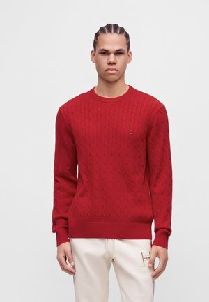 Jeune homme aux cheveux tressés porte un pull rouge torsadé et un pantalon blanc avec un petit logo sur la cuisse gauche, debout devant un fond uni.