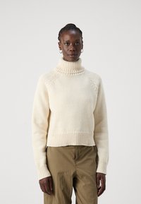 Cremefarvet turtleneck sweater med ribbede manchetter og kant, der har en hyggelig tekstur og løs pasform, parret med olivengrønne bukser.