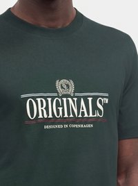 Σκούρο πράσινο μπλουζάκι με λευκό κείμενο "ORIGINALS" και μικρότερο κείμενο "DESIGNED IN COPENHAGEN" στο στήθος.