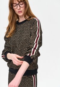 Zwarte en bruine leopardprint trui met zwarte geribbelde manchetten en een effen zwarte tailleband. Heeft bordeauxrode en witte gestreepte accenten op de mouwen.