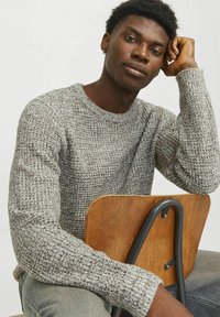 Jersey gris de punto con un patrón texturizado, cuello redondo y mangas largas. Modelo sentado en una silla de madera con un marco de metal.