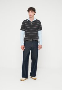 Μαύρο ριγωτό πόλο πουκάμισο πάνω από ένα ανοιχτό μπλε μπλουζάκι με μακριά μανίκια, συνδυασμένο με σκούρο μπλε τζιν παντελόνι με φαρδιά πόδια και μπεζ παπούτσια. Λεπτή μαύρη ζώνη με κύκλους.