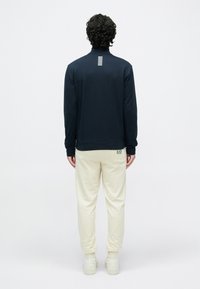 Tmavomodrá mikina s vysokým golierom, detailom pruhov na chrbte, v kombinácii s béžovými teplákmi a bielymi teniskami. Hladká textúra a voľný strih.