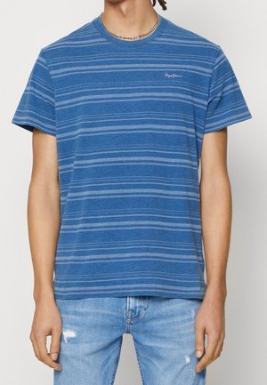 Homme portant un t-shirt rayé bleu à manches courtes et un jean bleu clair usé, raccourci au niveau du cou et des cuisses, debout, les bras détendus.