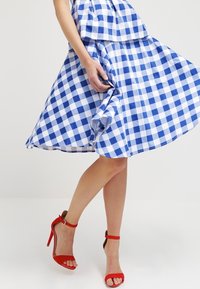 Blau-weiß kariertes Kleid mit schwingendem Rock und gestuftem Design. Kombiniert mit roten High-Heel-Sandalen. Glatte Stoffoberfläche.