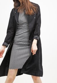 Svart mockatrenchcoat över en svartvit klänning med zickzackmönster, med en figurnära siluett och långa ärmar.