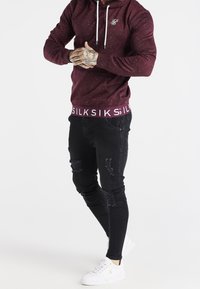 Uomo che indossa una felpa con cappuccio color bordeaux, jeans skinny neri strappati e sneaker bianche, in piedi con le mani giunte all'altezza della vita su sfondo bianco.