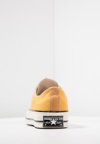 Gulld canvas sneakers med lav profil, hvit gummisåle og svart logo-patch på hælen. Har kontraststing langs baksiden.