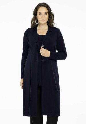 Marineblauwe lange cardigan van gladde stof met een kraag en knopen aan de voorkant. Gepaard met een bijpassende top en zwarte broek.