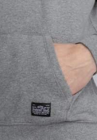 Sudadera gris hecha de tela suave, que cuenta con un bolsillo canguro y una etiqueta de marca negra con texto blanco en la esquina inferior izquierda.