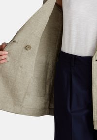 Svetle béžový texturovaný blazer s kostkovaným vzorem, na jedno knoflíček a se bočními kapsami, vrstvený přes tmavě modré kalhoty.