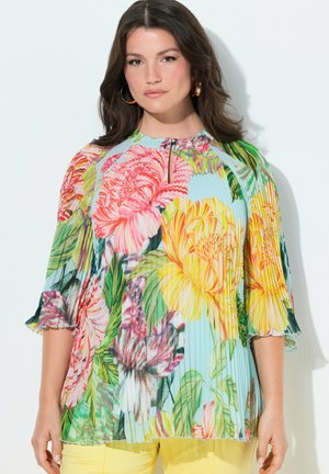 Kobieta o ciemnych włosach, nosząca złote kolczyki kółka i plisowaną kwiecistą bluzkę oraz żółte spodnie na białym tle.