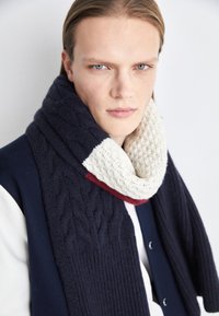 Navy cable-knit scarf featuring a cream textured band with red and cream accents. Thick, soft fabric with a cozy, layered design.