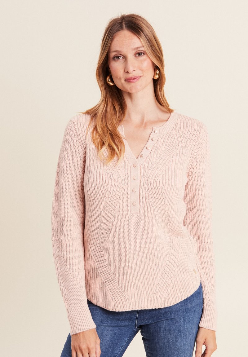 Helles rosa gestricktes Sweatshirt mit strukturiertem Muster, V-Ausschnitt mit drei Knöpfen und langen Ärmeln. Kombiniert mit blauen Jeans.