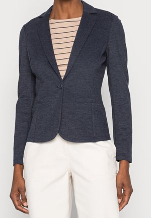 Nainen, jolla on päällään laivastonsininen istuva bleiseri beigen ja laivastonsinisen raidoitetun paidan päällä, yhdistettynä valkoisiin korkeavyötäröisiin housuihin.