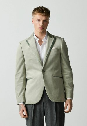 Young man with short blond hair wearing a light green velvet blazer, white dress shirt, and dark gray trousers against a plain background.